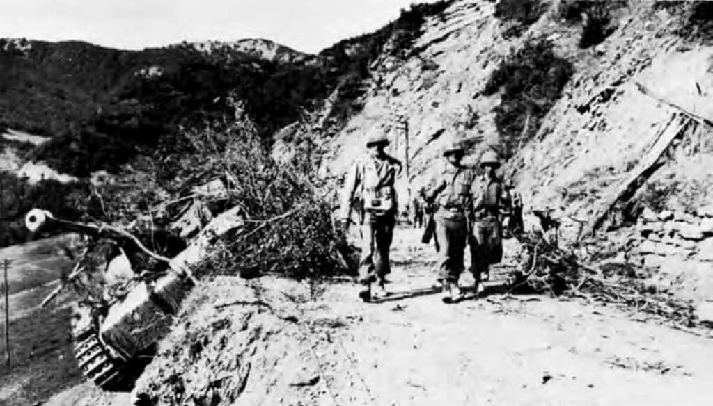 Tre soldati americani camminano su una strada sterrata di montagna; a sinistra, rovesciato sul ciglio, un mezzo corazzato con il lungo cannone.