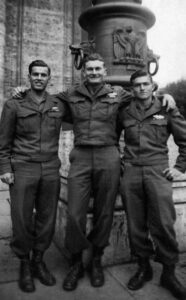 Tre soldati in uniforme militare degli anni '40, sorridenti e con braccia intorno alle spalle, davanti a un monumento storico.