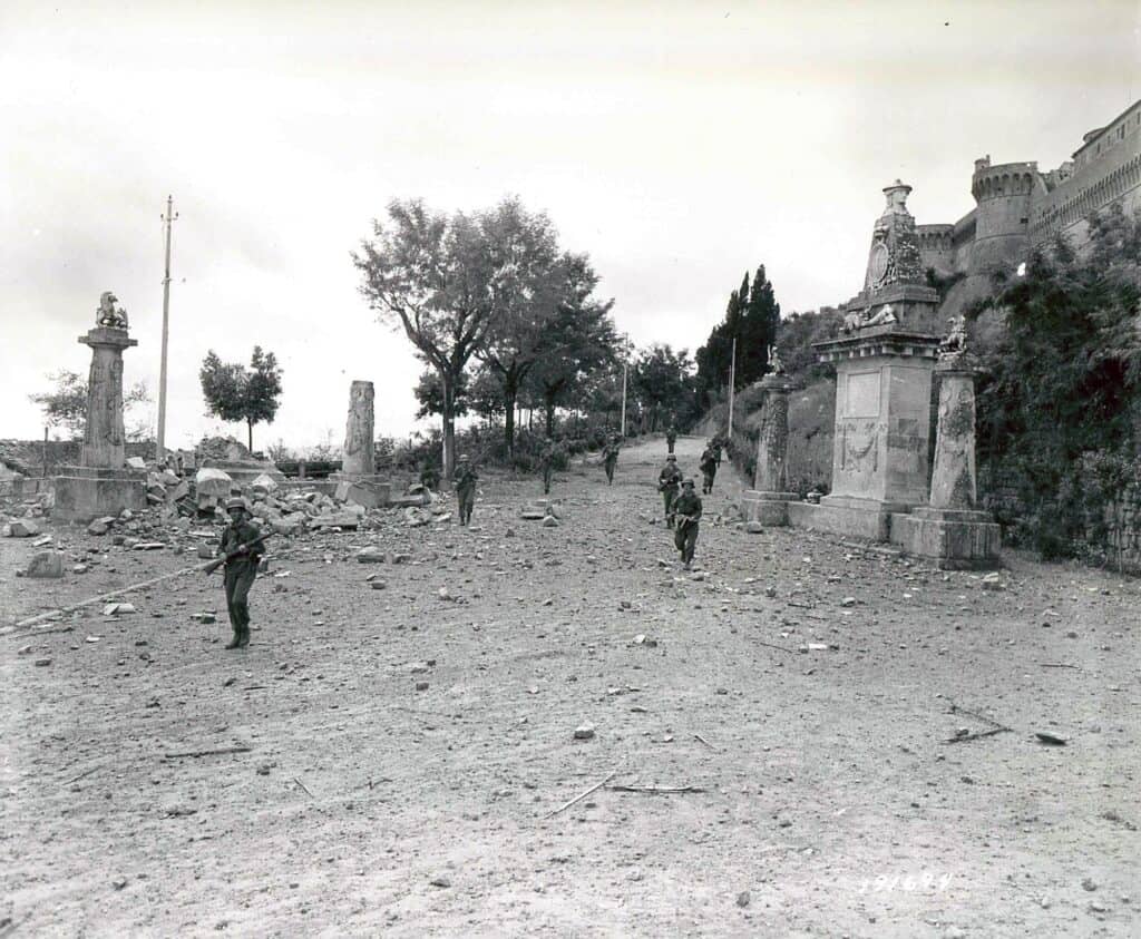Fotografia in bianco e nero: soldati americani camminano in fila, distanziati, su una strada coperta di macerie che sale verso le mura di Volterra; a destra un edificio merlato, ai lati pilastri in pietra.