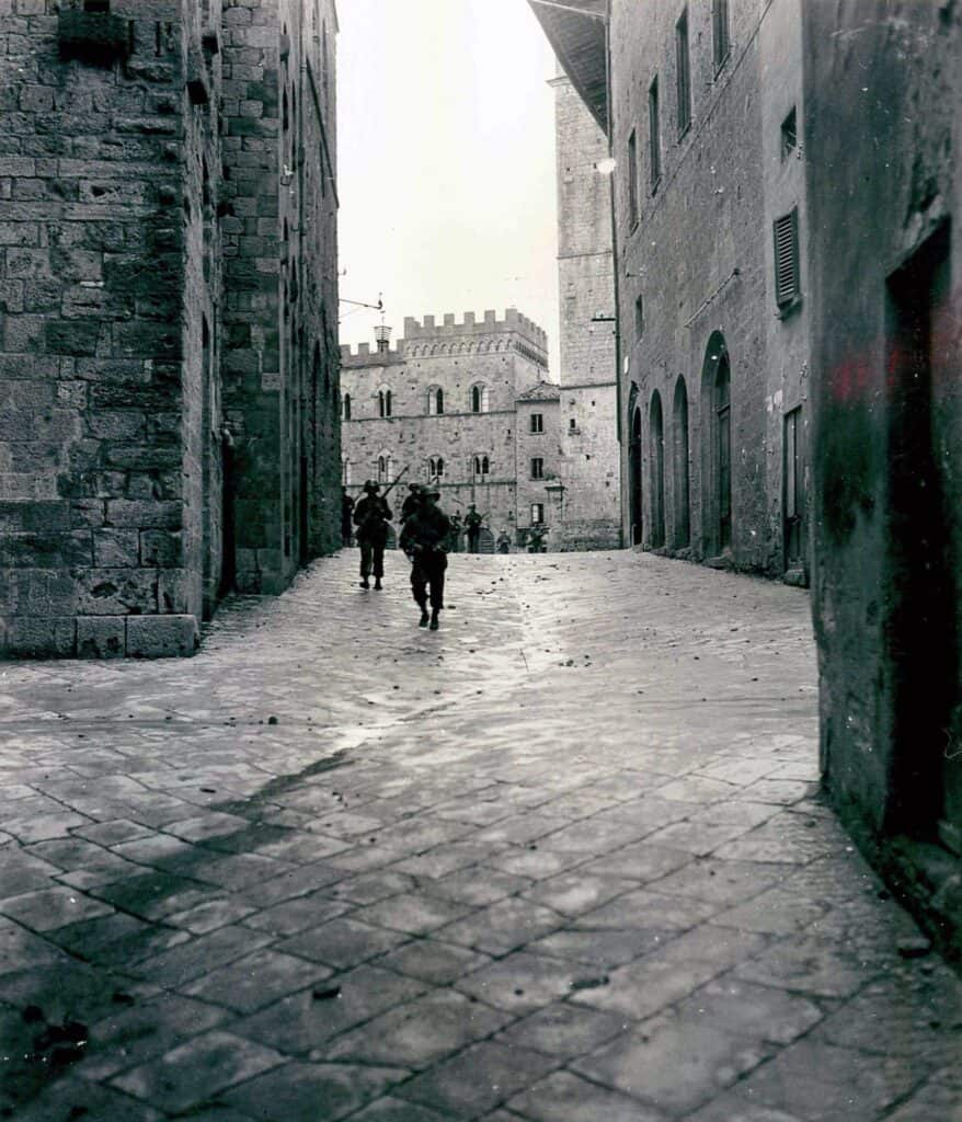 Fotografia in bianco e nero: due soldati americani camminano in una stretta via lastricata di Volterra, fra alti palazzi di pietra; in fondo un palazzo con una torre merlata.