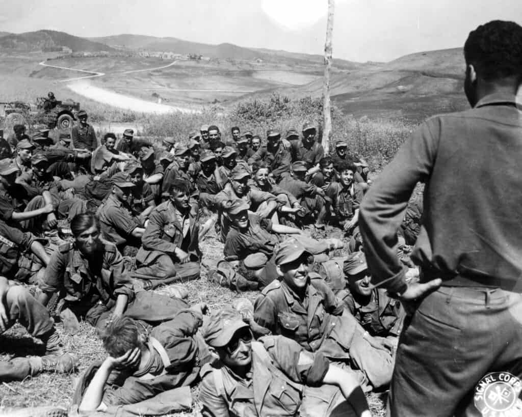 Fotografia in bianco e nero: decine di soldati tedeschi prigionieri seduti e sdraiati sull'erba di un pendio; un soldato americano di spalle parla con loro; colline sullo sfondo.