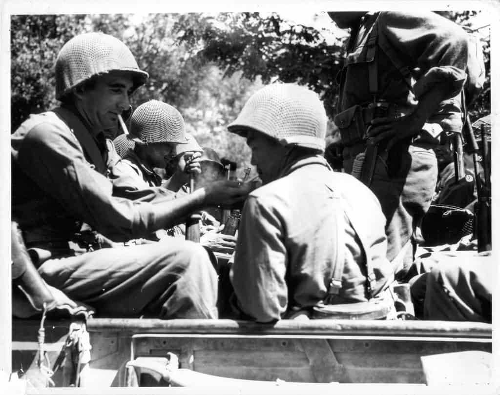 Fotografia in bianco e nero: due soldati americani con l'elmetto, seduti fra altri commilitoni su un mezzo militare, si accendono una sigaretta.