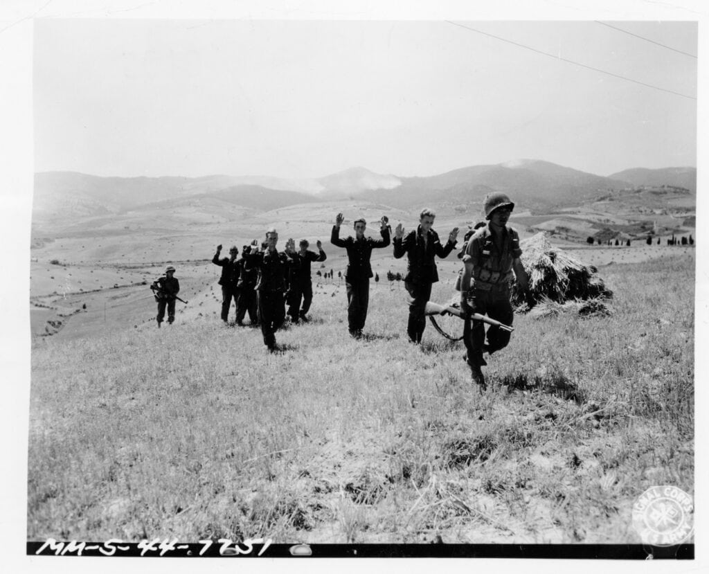 Fotografia in bianco e nero: su una collina con l'erba alta, alcuni soldati tedeschi camminano in fila con le mani alzate; un soldato americano li segue con il fucile; sullo sfondo colline.