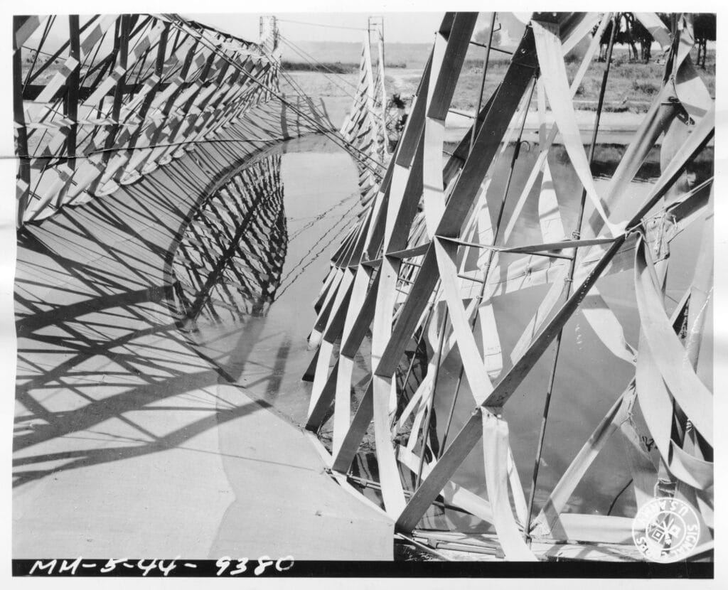 Fotografia in bianco e nero: vista ravvicinata del falso ponte sull'Arno, un'intelaiatura a traliccio di travi e listelli rivestita in parte di strisce di tela, con le ombre proiettate sull'acqua; sullo sfondo la riva con alberi.