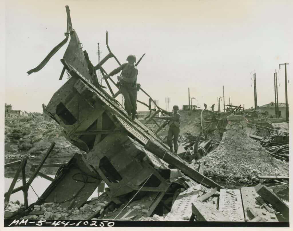 Fotografia in bianco e nero: due soldati americani con l'elmetto scavalcano le travi contorte e le macerie di un ponte ferroviario distrutto sull'Arno a Pisa; sullo sfondo pali e case.