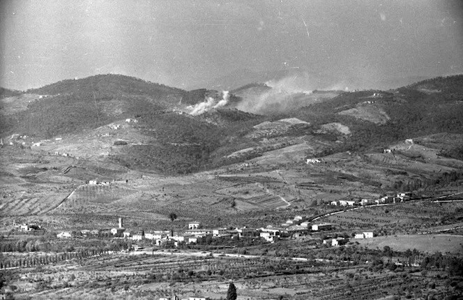 Veduta di colline toscane con colonne di fumo di esplosioni su una cresta; a sinistra il villaggio di San Michele.
