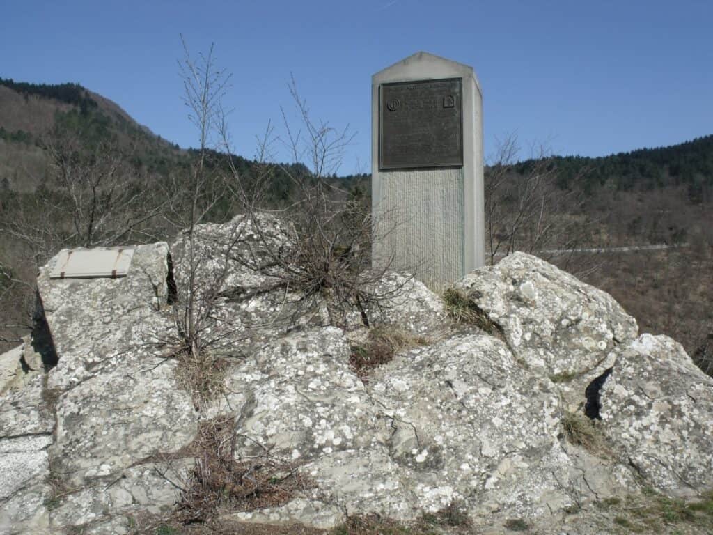 Un cippo di pietra con una targa di bronzo sorge su un cumulo di rocce, fra alberi spogli e colline.