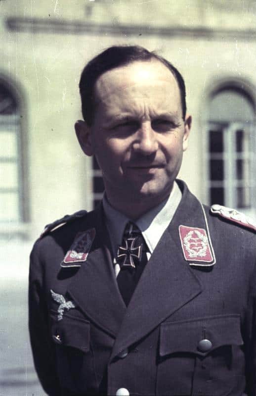 Ritratto a colori, a mezzo busto, di un ufficiale della Luftwaffe in uniforme grigio-azzurra, con mostrine rosse sul colletto e una croce di ferro al collo, davanti a un edificio.