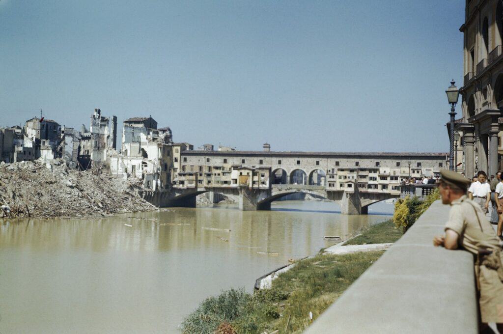 Fotografia a colori: il Ponte Vecchio sull'Arno, con le sue botteghe, visto dalla riva nord. A sinistra, sulla riva opposta, cumuli di macerie e case sventrate. A destra un porticato e alcuni civili raccolti vicino all'imbocco del ponte; in primo piano un militare in uniforme kaki e berretto a visiera, appoggiato al parapetto del lungarno.